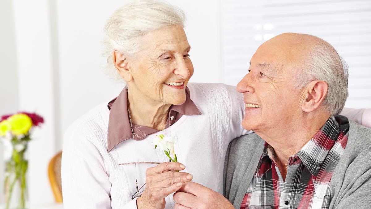 An eldery couple smiling at one another holding flowers