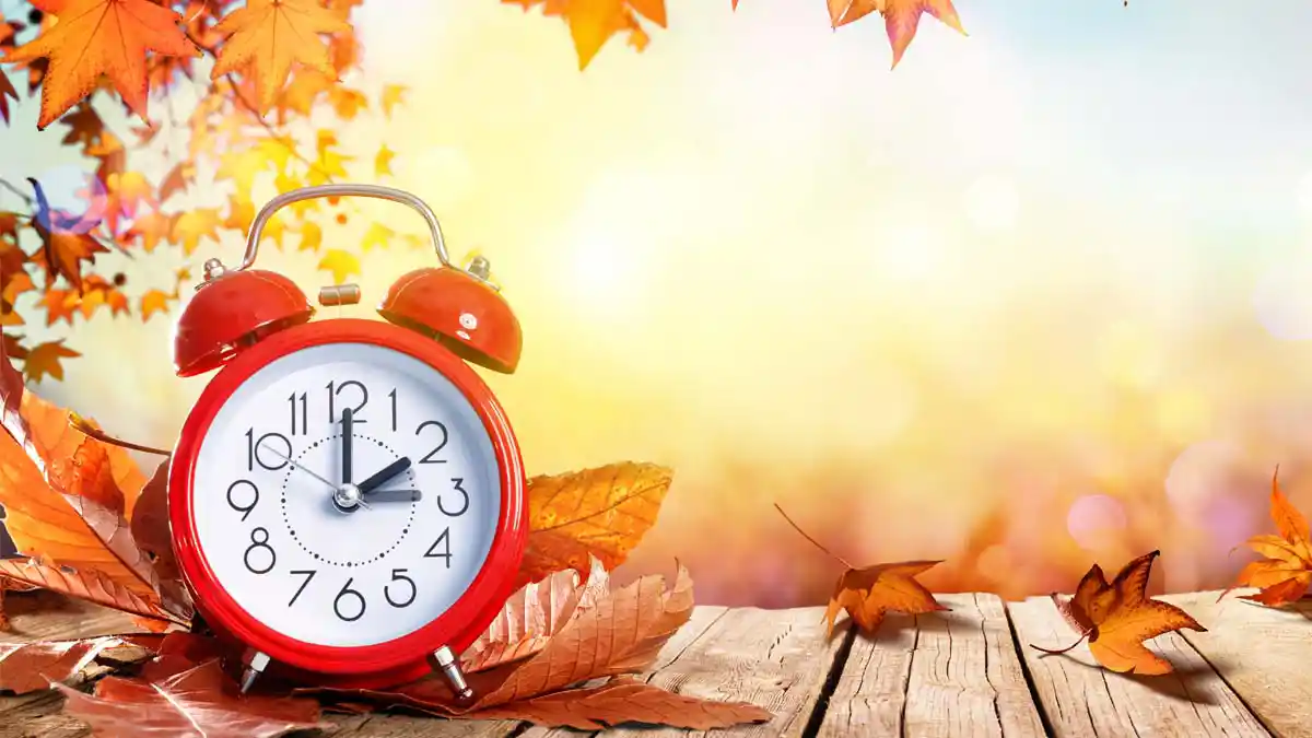 An alarm clock with the sun shining and fall leaves in the background reminds us about Daylight Saving Time